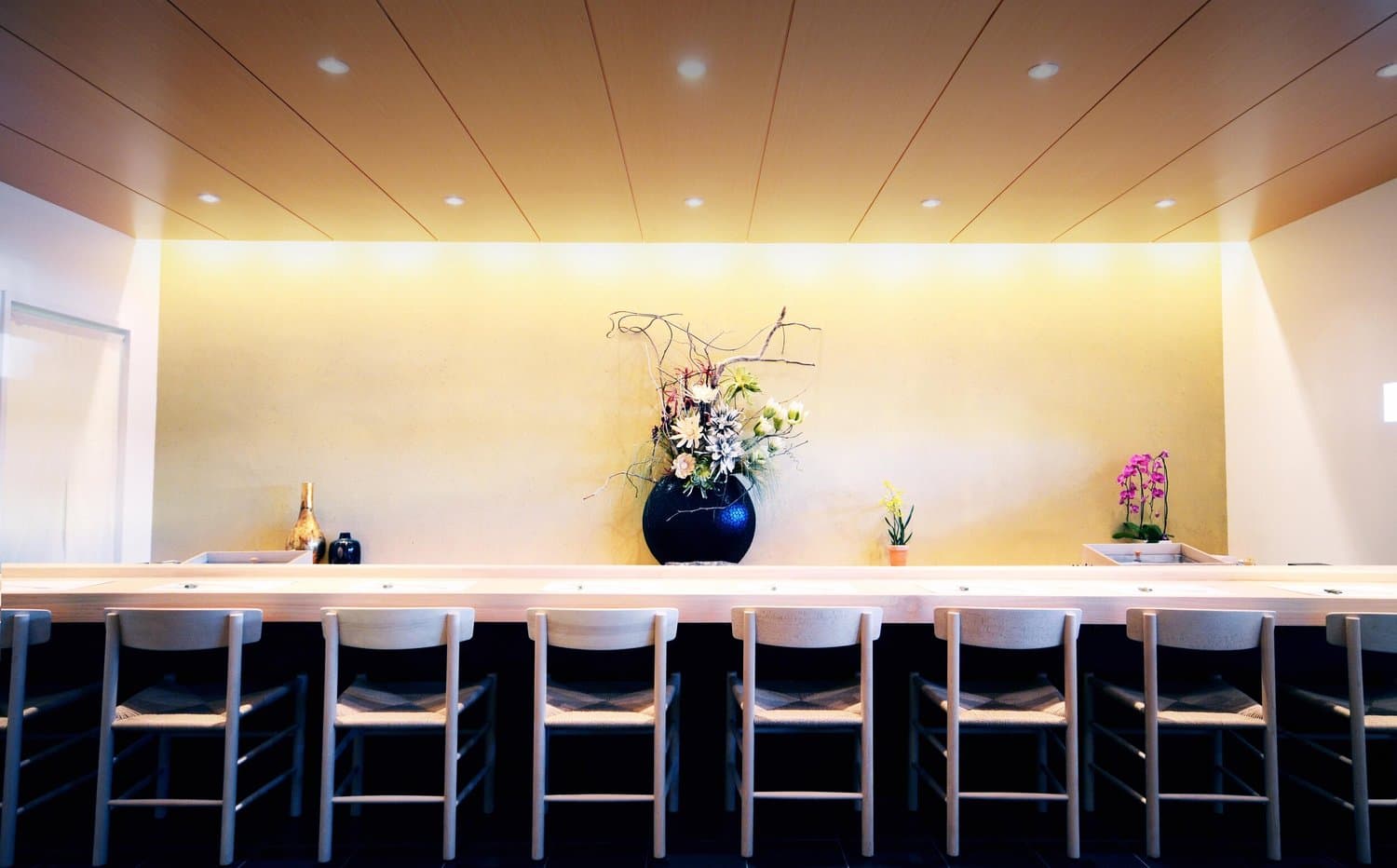 Modern sushi bar counter with white seating, illuminated yellow wall, and floral arrangement