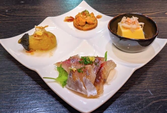 Plated appetizers with shrimp dumplings, sliced fish, and egg custard in black bowl