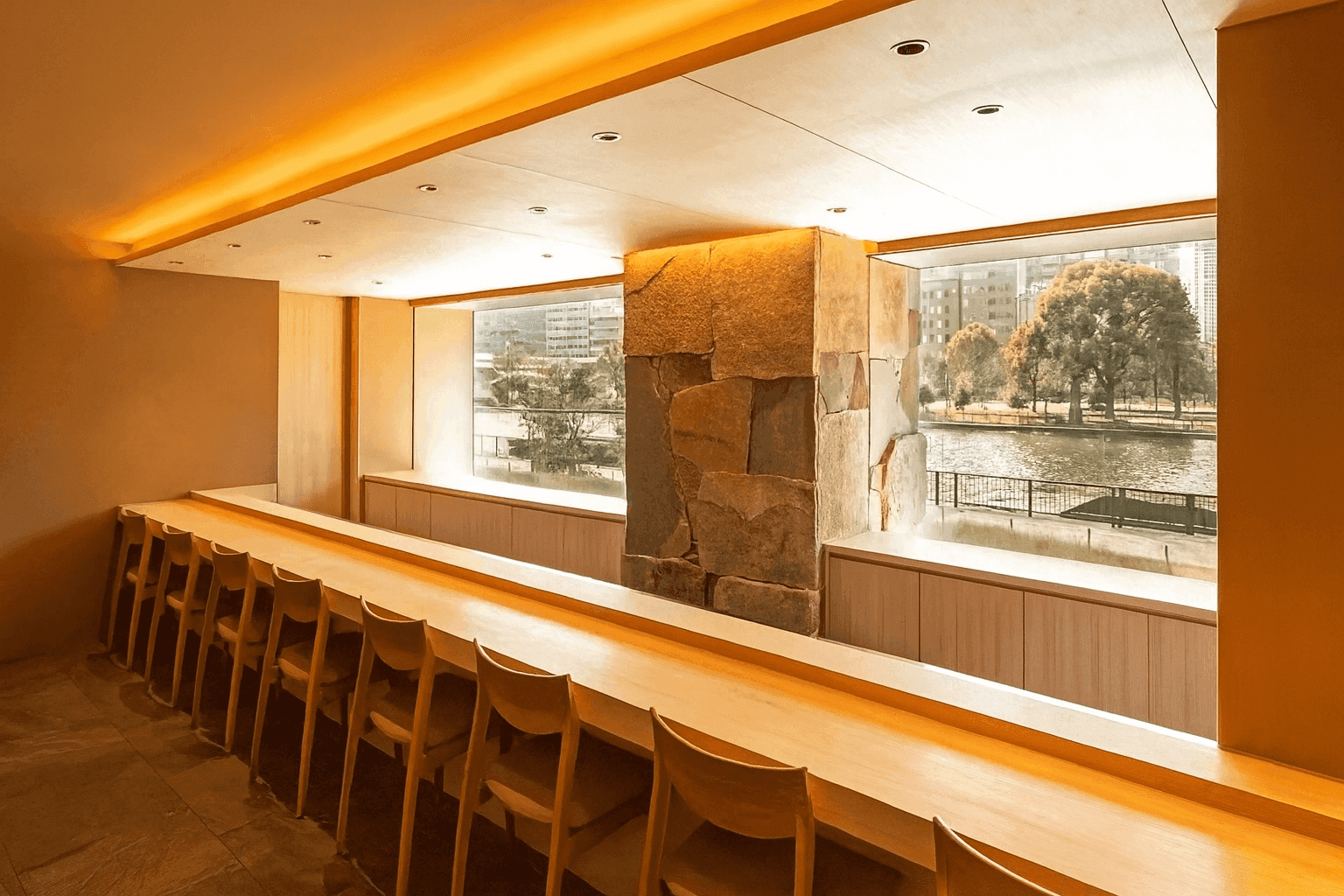 Modern sushi bar with wooden counter, stone accent wall, and river views through floor-to-ceiling windows