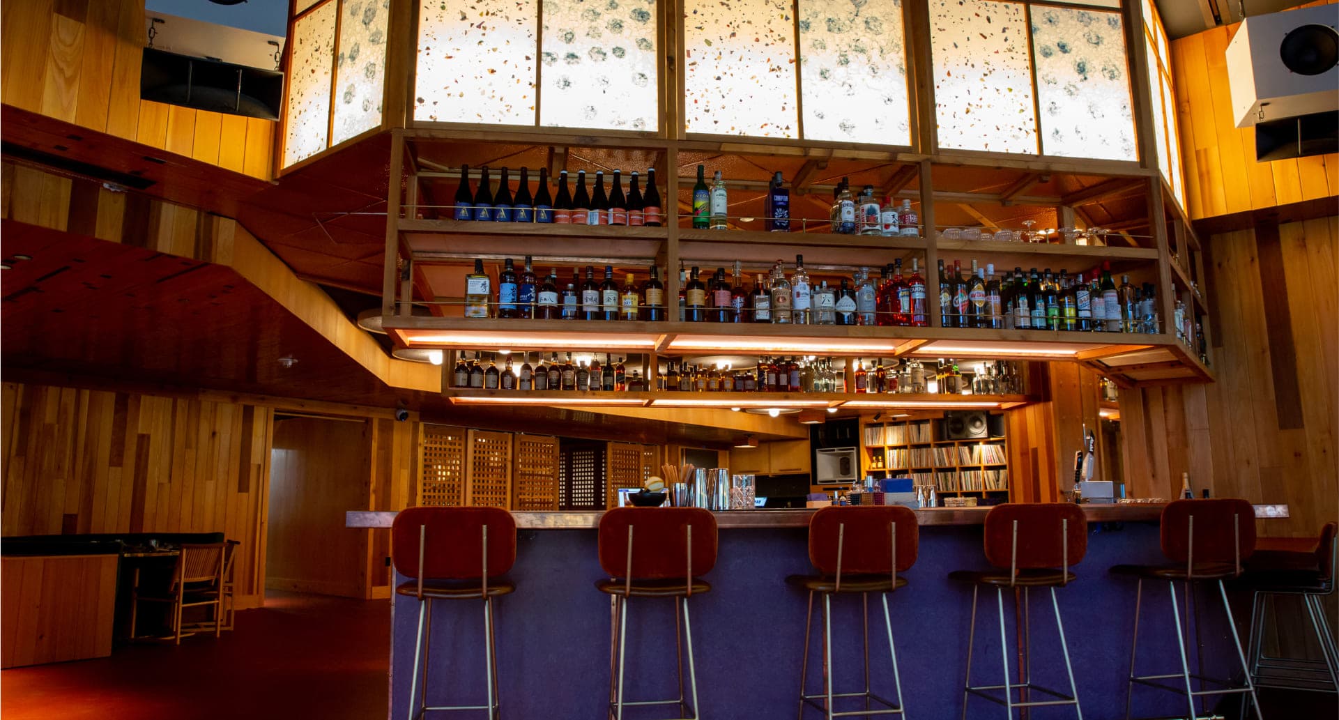 Modern bar with wooden interior, shelves of spirits, and burgundy bar stools under skylights