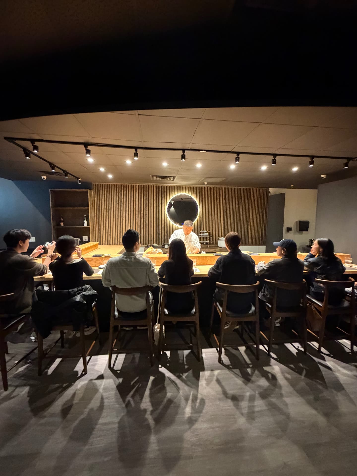 Sushi preparation counter with chef and guests seated at wooden bar