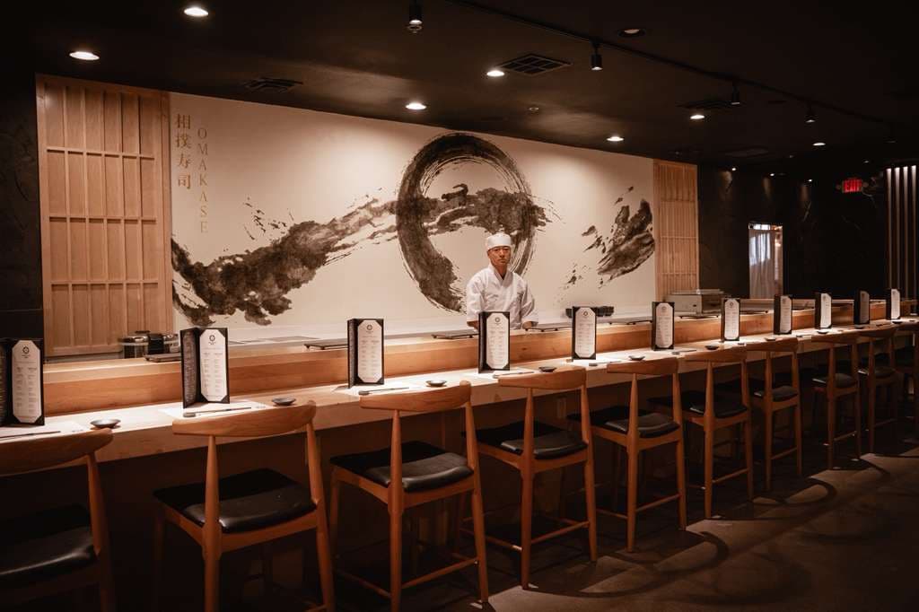 Sushi chef in white uniform standing behind wooden counter with artistic mural backdrop